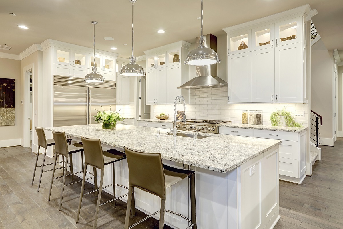 White kitchen design features large bar style kitchen island with granite countertop illuminated by modern pendant lights. Stainless steel appliances framed by white shaker cabinets . Northwest USA